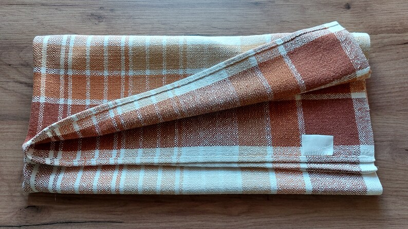 May include: Folded plaid kitchen towel in shades of brown, orange, and cream. The woven fabric features a classic check pattern. A small, blank white tag is sewn on the towel. The towel is folded neatly.