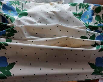 May include: A white tablecloth with a green and blue floral pattern and black polka dots. The tablecloth is folded in the middle and has a slight wrinkle.