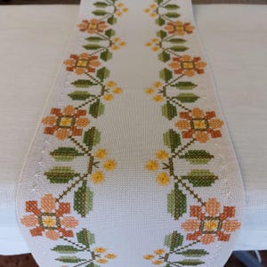 Puede incluir: Un camino de mesa de lino blanco con diseños florales bordados en tonos naranja, amarillo y verde. Las flores están dispuestas en un patrón repetitivo a lo largo de los bordes del camino de mesa.