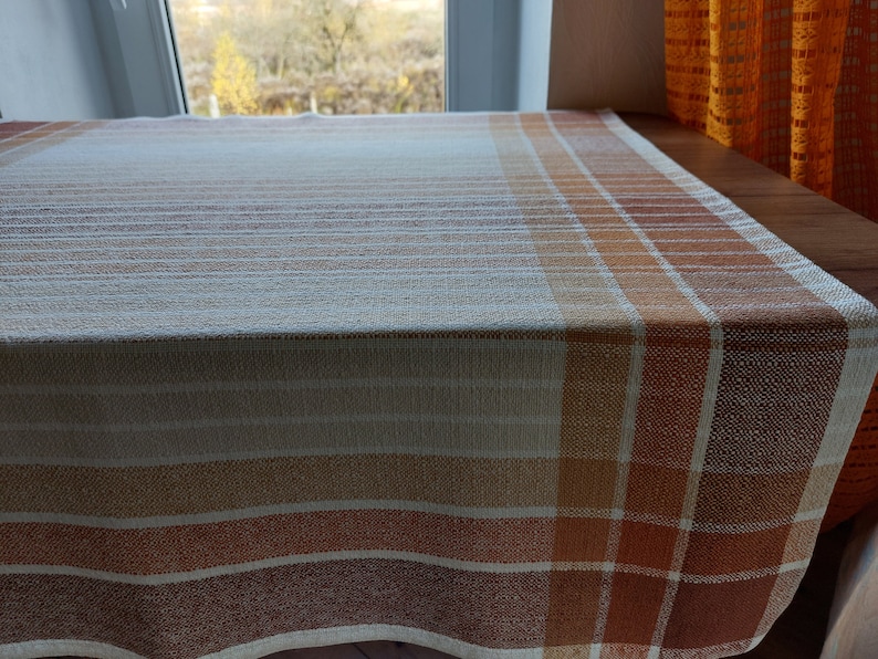 May include: A rectangular tablecloth with a plaid pattern in shades of white, beige, orange, and brown. The fabric has a woven texture. The tablecloth is draped over a wooden table, with a window and orange curtain in the background.