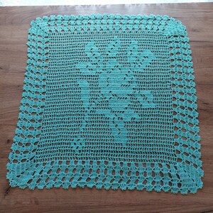 May include: A square, turquoise crocheted doily with a floral design in the center. The edges feature a scalloped pattern. The doily is made of a soft, textured yarn and is displayed on a wooden surface.