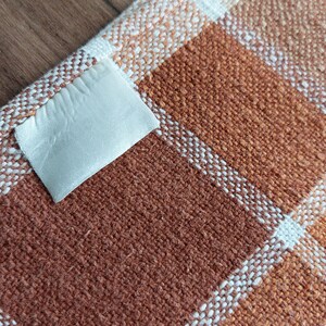 May include: Close-up of a woven fabric with a plaid pattern in shades of brown, orange, and white. A small, off-white label is attached to the fabric. The fabric appears to be a textile, possibly for home decor or apparel.