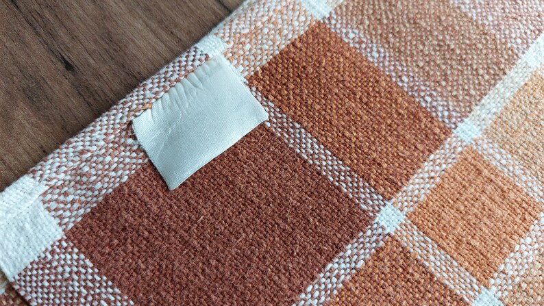 May include: Close-up of a woven fabric with a plaid pattern in shades of brown, orange, and white. A small, off-white label is attached to the fabric. The fabric appears to be a textile, possibly for home decor or apparel.