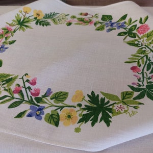 May include: White linen tablecloth with a green, yellow, pink, and blue floral border. The border features a variety of flowers and leaves, including clover, violets, and sweet peas. The tablecloth has a "Jolis" signature in the lower right corner.