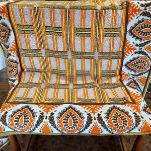 May include: A colorful tablecloth with a geometric pattern in orange, white, and brown. The design features vertical stripes and rectangular blocks, with decorative floral borders. The fabric is draped over a wooden table.