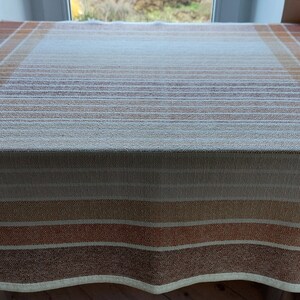 May include: A rectangular tablecloth with a plaid and striped pattern in shades of brown, orange, and cream. The fabric is laid out on a wooden surface, with the window in the background.