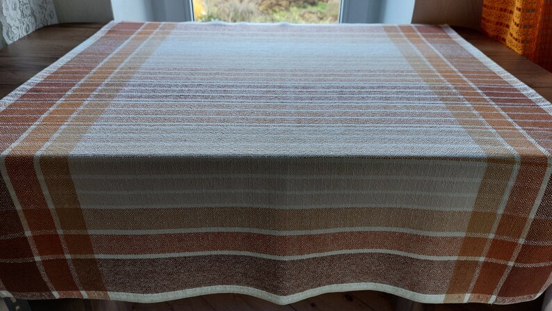 May include: A rectangular tablecloth with a plaid and striped pattern in shades of brown, orange, and cream. The fabric is laid out on a wooden surface, with the window in the background.