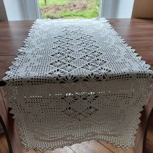 May include: White crocheted table runner with a delicate floral pattern. The runner has a scalloped edge and is made of a fine, lightweight yarn.