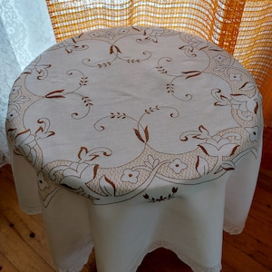 May include: White tablecloth with a brown floral embroidery design. The tablecloth has a lace trim around the edges.