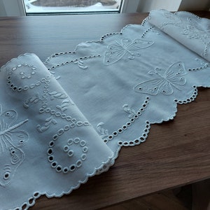 Puede incluir: Camino de mesa blanco bordado con un borde festoneado y un diseño de mariposa. El camino de mesa está hecho de tela delicada y presenta detalles intrincados.