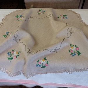 May include: A beige round tablecloth with a crocheted edge and embroidered floral designs. The tablecloth features a central square design with a scalloped edge and four corner designs.
