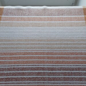 May include: A rectangular, woven textile with a plaid design. The color palette includes shades of brown, orange, and white, creating a warm, inviting aesthetic. The textile features both horizontal stripes and vertical lines, forming a grid pattern.