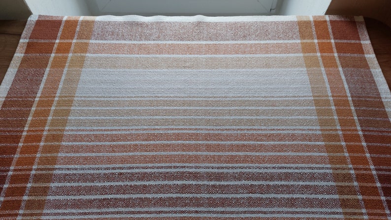 May include: A rectangular, woven textile with a plaid design. The color palette includes shades of brown, orange, and white, creating a warm, inviting aesthetic. The textile features both horizontal stripes and vertical lines, forming a grid pattern.