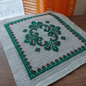 May include: A square, beige linen placemat with a green and brown embroidered floral design.