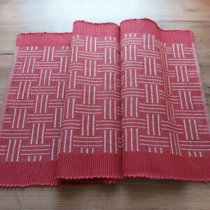 May include: Red woven placemats with a white geometric pattern. The placemats are folded in thirds and laid out on a wooden surface.