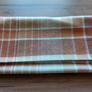 May include: Folded plaid kitchen towel in shades of brown, orange, and cream. The woven fabric features a classic checkered pattern. The towel is folded neatly, showcasing the pattern and texture of the material.