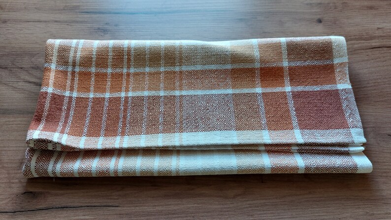 May include: Folded plaid kitchen towel in shades of brown, orange, and cream. The woven fabric features a classic checkered pattern. The towel is folded neatly, showcasing the pattern and texture of the material.
