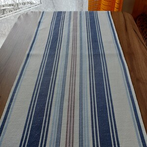 May include: A white and blue striped table runner with a subtle red and blue stripe pattern. The runner is made of a woven fabric and has a slightly textured surface.