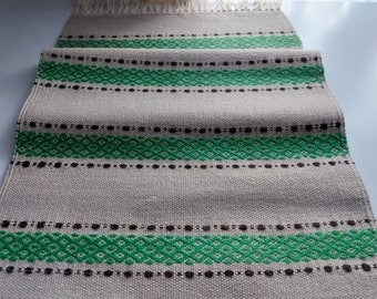 May include: A beige table runner with green and brown woven stripes. The runner has a fringe on the ends.