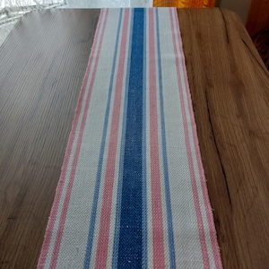 May include: A white table runner with blue and pink stripes. The runner is made of a woven fabric and has a slightly textured surface.