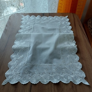 May include: A white rectangular table runner with a scalloped edge and intricate floral embroidery. The runner is made of a soft, light-colored fabric and is placed on a wooden table. The design features delicate cutwork and a classic, elegant style.