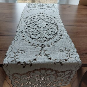 May include: White crocheted table runner with a delicate floral pattern. The runner features a large oval design in the center and smaller floral motifs along the edges.