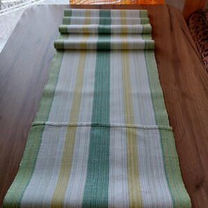May include: A green, yellow, and white striped table runner. The runner is made of woven fabric and is folded over at the top. The stripes run vertically down the length of the runner.