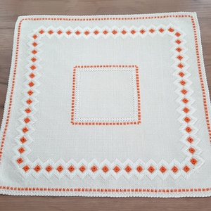 Può includere: Un runner da tavolo in lino bianco con un bordo ricamato arancione. Il bordo presenta un motivo a rombi con piccoli quadrati arancioni.