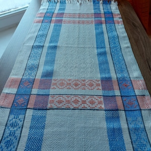 May include: A white linen table runner with blue and pink stripes and a woven pattern. The runner has a fringed edge.