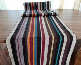 May include: A woven table runner with vertical stripes in various colors, including black, brown, teal, and orange. The runner is rolled up at one end, showcasing the striped pattern. The runner is placed on a wooden table.