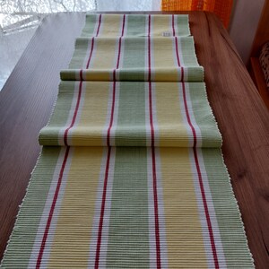 May include: A striped table runner in shades of yellow, green, red, and white. The runner is made of a textured fabric and is laid out on a wooden table. The stripes run lengthwise, creating a simple yet elegant design.