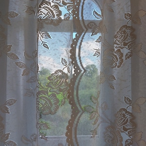 Puede incluir: Cortinas blancas de encaje con un patrón floral cuelgan en una ventana, permitiendo una vista del follaje verde afuera.