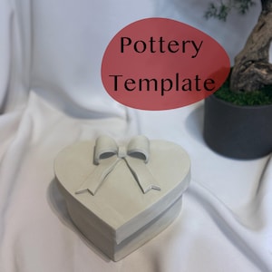 May include: A heart-shaped pottery box with a bow on the lid. The box is light gray and sits on a white surface. The text "Pottery Template" is in a red oval in the background. A small potted plant is also in the background.
