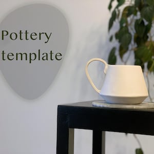 Op de afbeelding: Witte keramische mok met een afgeronde handgreep, die op een zwarte tafel staat met een grijze onderzetter. De mok heeft een unieke, licht afgeschuinde vorm. De tekst "Pottery template" is zichtbaar op de achtergrond.