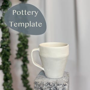 Puede incluir: Una taza de cerámica blanca con asa, sentada sobre una superficie de piedra gris. La taza tiene una superficie ligeramente texturizada y una forma redondeada. El texto "Pottery Template" es visible en el fondo.