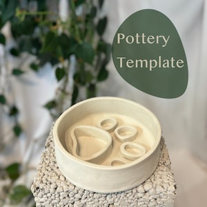 Puede incluir: Una plantilla de cerámica beige claro con un diseño de huella de pata. La plantilla tiene forma circular con una serie de recortes ovalados y en forma de corazón. El texto "Pottery Template" está en un óvalo verde en el fondo. La plantilla está sobre una superficie texturizada gris claro.