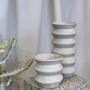 Może przedstawiać: Dwa szare ceramiczne wazony z żebrowanym wzorem. Wazony stoją na szarej, fakturowanej powierzchni.