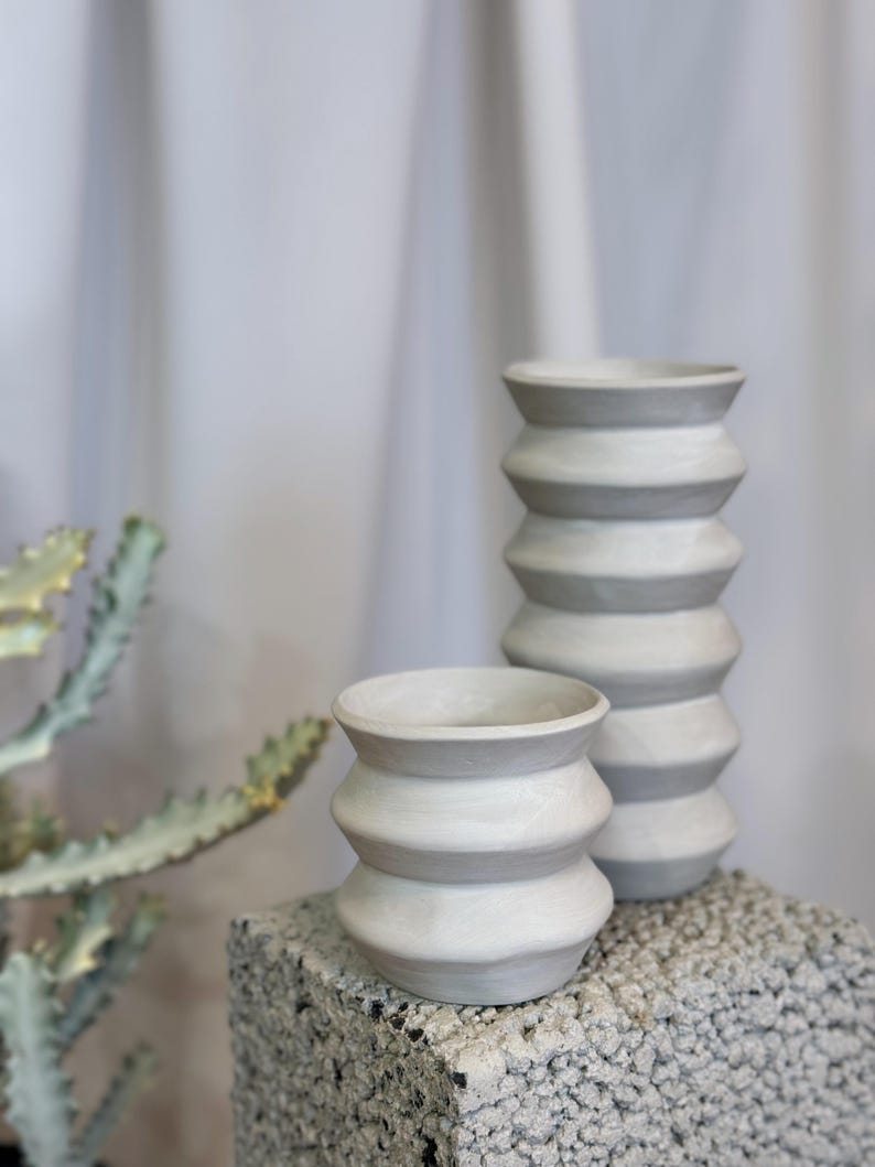 Może przedstawiać: Dwa szare ceramiczne wazony z żebrowanym wzorem. Wazony stoją na szarej, fakturowanej powierzchni.