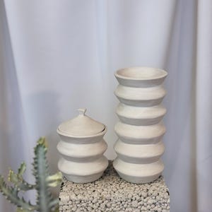 Może przedstawiać: Dwa białe ceramiczne pojemniki o fakturowanym, falistym wzorze. Większy pojemnik to wazon, a mniejszy pojemnik ma pokrywkę.