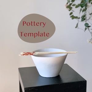May include: A white ceramic bowl with a rounded bottom and a wide rim, sitting on a black surface. A pair of wooden chopsticks with red accents are resting on the rim of the bowl.