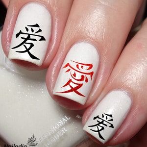 May include: Close-up of a hand with white nail polish and red and black nail art featuring the Japanese kanji character for "love" (愛).