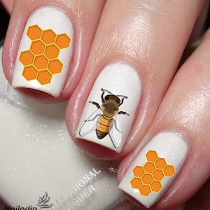 May include: White nail polish with a bee and honeycomb design. The bee is brown and yellow with black stripes. The honeycomb is orange and yellow.