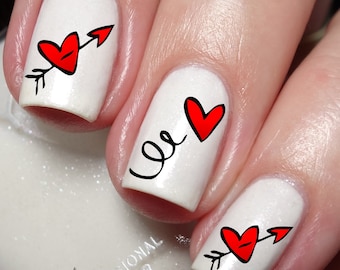 Pode incluir: Esmalte de unha branco com desenhos de coração e flecha vermelhos. A arte de unha apresenta um coração com uma flecha atravessando-o, um coração com um redemoinho e um coração com uma flecha.