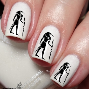 May include: Three white fingernails with black nail art of an Egyptian god with a bird head and a staff. The nail art is on the index, middle, and ring fingers.
