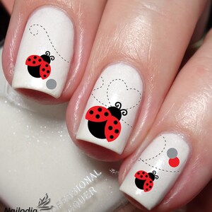 May include: White nail polish with ladybug nail art decals. The ladybugs are red with black spots and have black wings. The ladybugs are flying with a dotted line trail.
