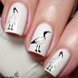 May include: White nail polish with black ibis bird nail art decals. The decals are on four fingernails. The nail polish bottle is visible in the bottom left corner of the image.
