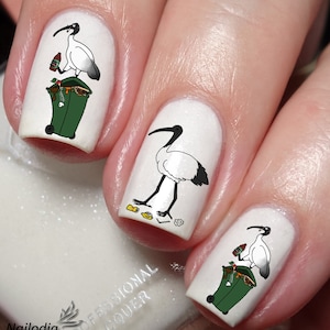 May include: White painted fingernails with a design of a white ibis bird standing next to a trash can with garbage on the ground. Another fingernail has a design of a white ibis bird standing on top of a green trash can with garbage inside. The text "Nailodia Professional Nail Lacquer" is visible on the bottom left corner of the image.