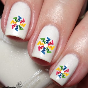 Peut inclure: Vernis à ongles blanc avec un motif géométrique coloré sur chaque ongle. Le motif est un cercle avec un motif en étoile en rouge, jaune, vert et bleu.