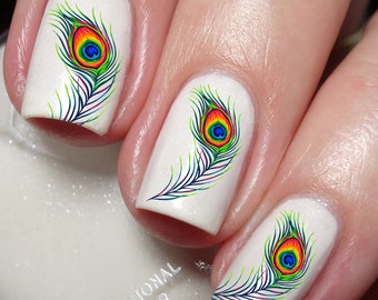 May include: Three fingernails painted with a solid white base coat, each featuring a vibrant, multi-colored peacock feather decal. The feathers display shades of blue, green, yellow, and red. The text Nailodia is visible at the bottom left.
