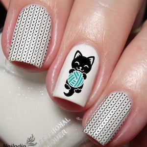 May include: Close-up of a hand with white nail polish and black and white knit nail art. One nail features a black cat holding a teal ball of yarn.